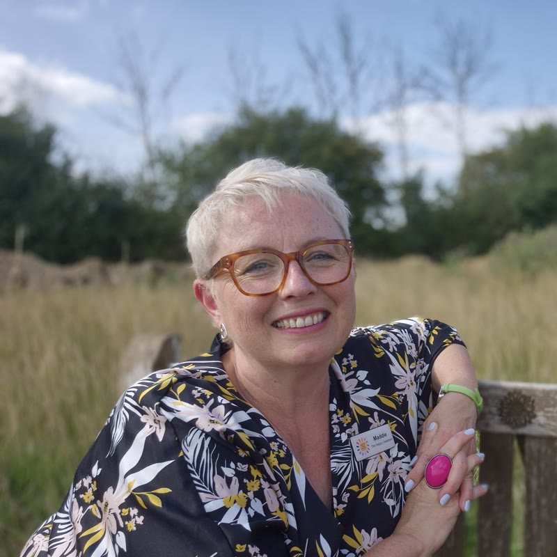 Maddie The Happy Celebrant, smiling, leaning on a wooden bench in a meadow